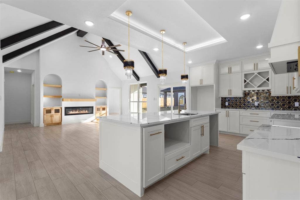 Vaulted ceilings with exposed beams, built-in fireplace with shelving, kitchen island with built-in sink, white cabinetry with gold-tone hardware, and a hexagonal tile backsplash