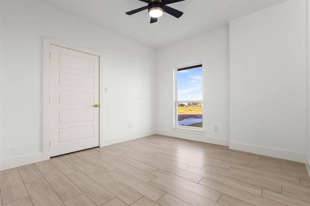 Wood-finish flooring, white walls, five-panel interior door with brass hardware, ceiling fan with integrated lighting, and a vertical window