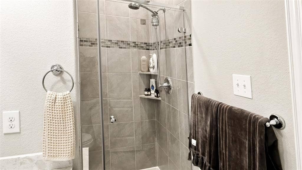 Walk-in shower featuring glass door enclosure, neutral-tone wall tiling, decorative horizontal tile inlay, integrated corner shelving, and a handheld shower fixture