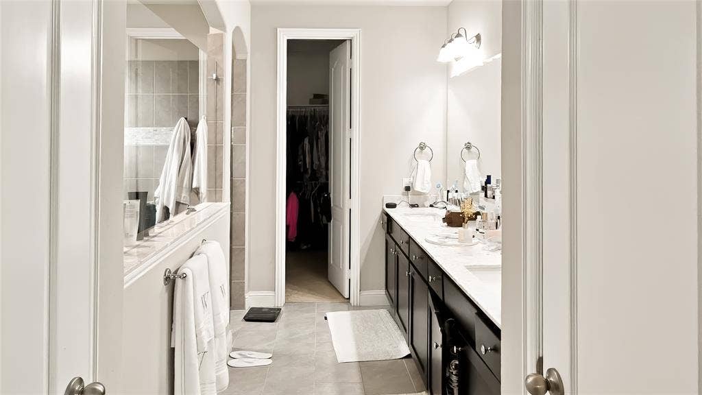Dual-sink vanity with dark cabinetry, light stone-surface countertops, tile flooring, and walk-in closet with built-in shelving