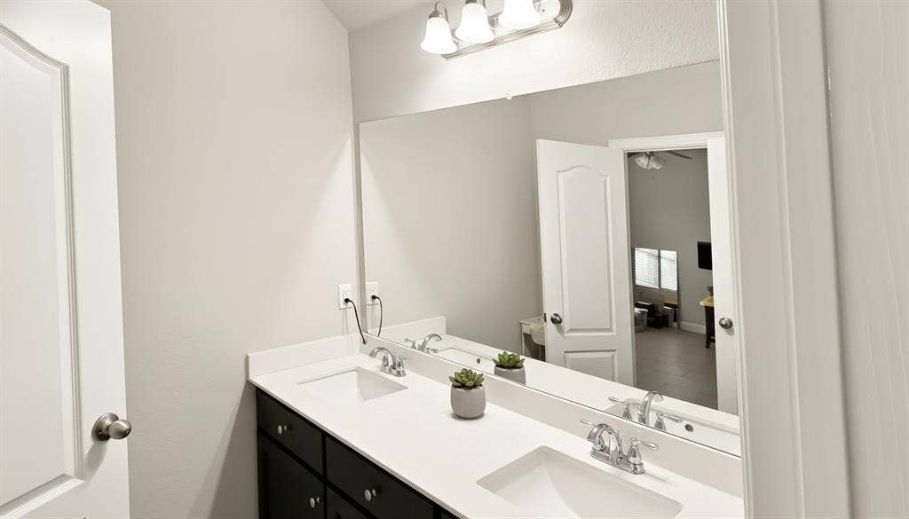 Double vanity featuring dark cabinetry, solid-surface countertop, dual integrated rectangular sinks, polished chrome faucets, and full-width vanity mirror with integrated three-bulb light fixture