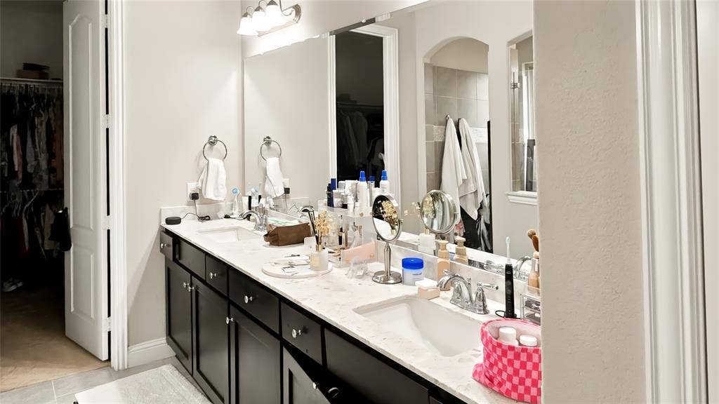 Double vanity featuring dark cabinetry, dual undermount sinks, light-toned stone-finish countertops, and a full-width vanity mirror with integrated lighting