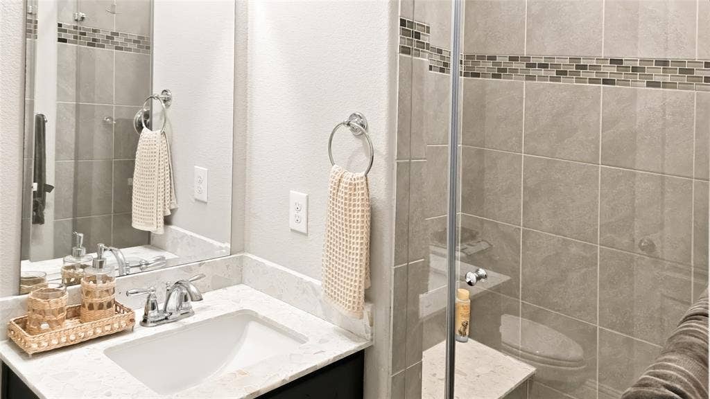 Bathroom featuring a vanity with a white composite countertop, an undermount rectangular sink, chrome hardware, a frameless glass shower enclosure with floor-to-ceiling tile, and a decorative tile accent inlay