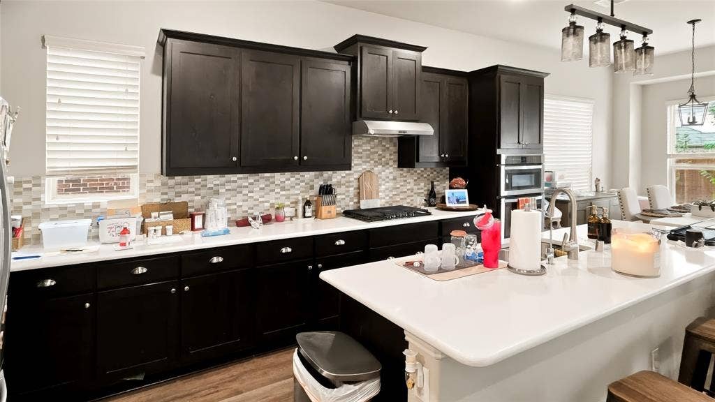 Kitchen featuring dark-finish cabinetry, white countertops, mosaic tile backsplash, built-in double wall ovens, and a center island with sink