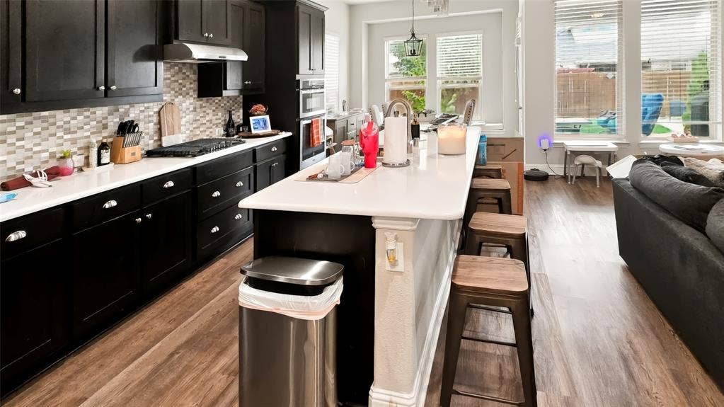 Kitchen featuring dark cabinetry, expansive white countertops, a tiled backsplash, built-in stainless steel double ovens, and wood-finish flooring