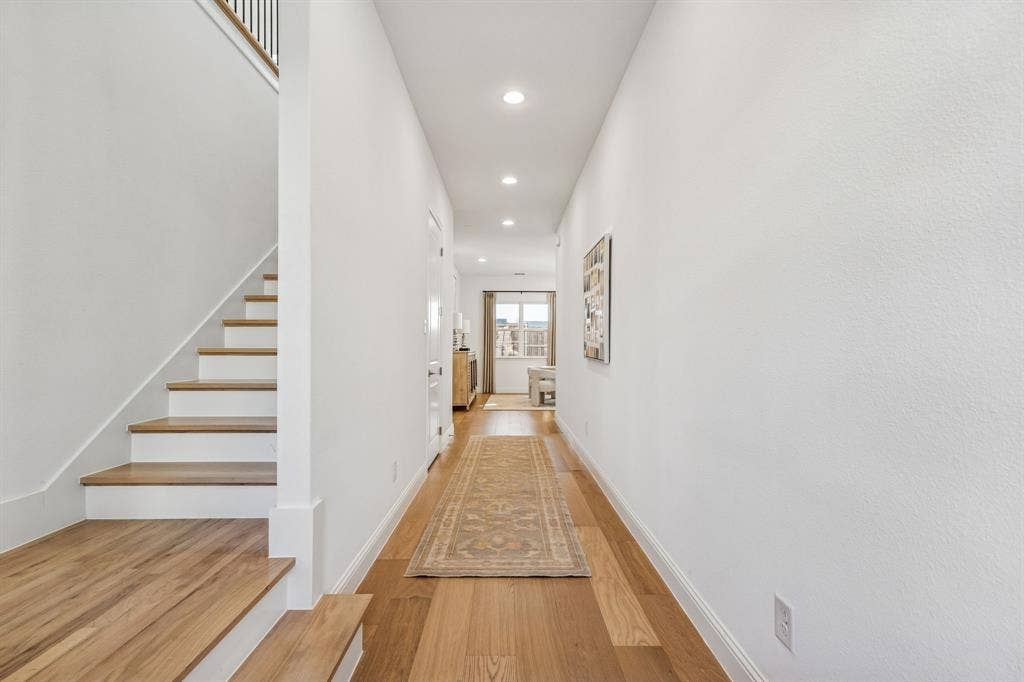 Wood-finish flooring throughout, white painted walls, recessed lighting, and a staircase with wood-finish treads and a dark metal railing