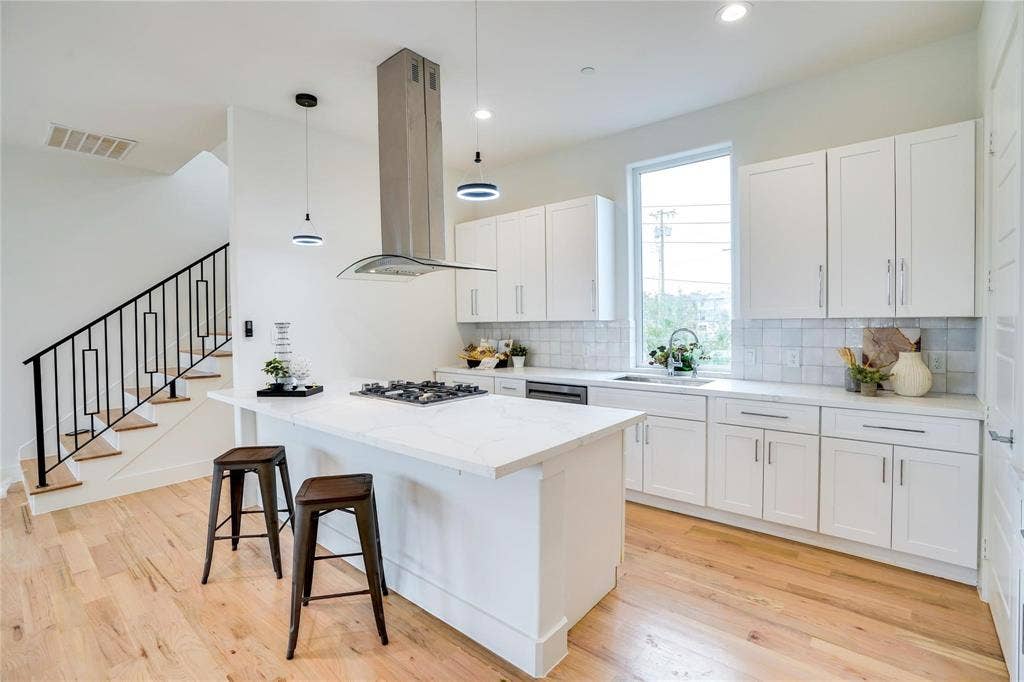 1612 Bennett Avenue, #105, Dallas, TX 75206 Contemporary kitchen featuring a center island with a built-in gas cooktop, white cabinetry, light-toned wood-finish flooring, a stainless steel range hood, and a full-height window