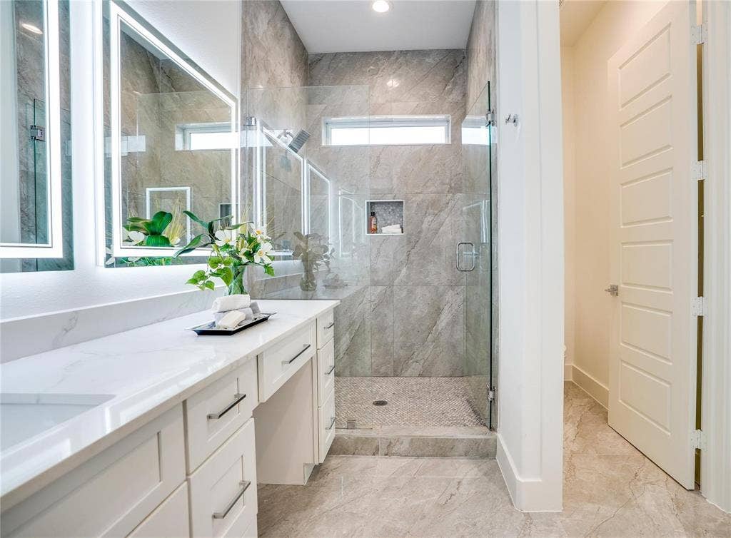 1612 Bennett Avenue, #105, Dallas, TX 75206 Modern bathroom featuring a double vanity with stone countertops and undermount sinks, frameless glass shower enclosure, large format tile flooring, and illuminated mirrors