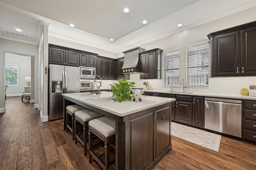 6168 Sweeney Trail, Frisco, TX 75034 Kitchen featuring wood-finish flooring, dark wood cabinetry, stainless steel appliances, a center island with a light-toned countertop, and recessed lighting