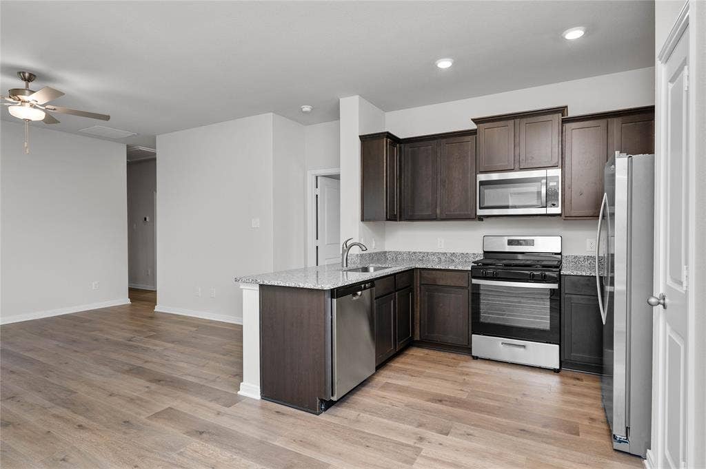 Kitchen with appliances with stainless steel finishes, dark brown cabinets, a peninsula, light wood-style floors, and light stone counters