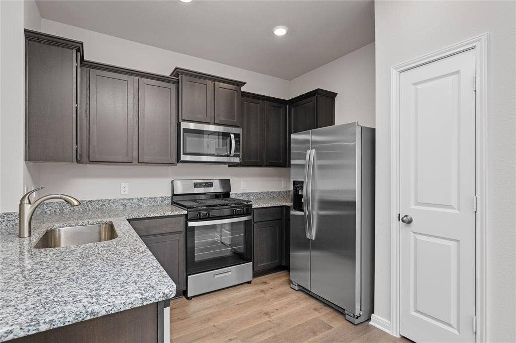 Kitchen featuring appliances with stainless steel finishes, light stone countertops, light wood-style flooring, and dark brown cabinets