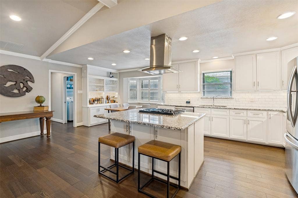 9421 Meadowknoll Drive, Dallas, TX 75243 Kitchen featuring a central island with a gas cooktop and granite countertop, stainless steel range hood, white shaker-style cabinetry, subway tile backsplash, and wood-finish flooring