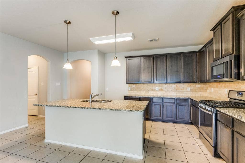 Kitchen featuring an island with an undermount sink, pendant lighting, dark wood-finish cabinetry, stainless steel appliances, and a tile backsplash