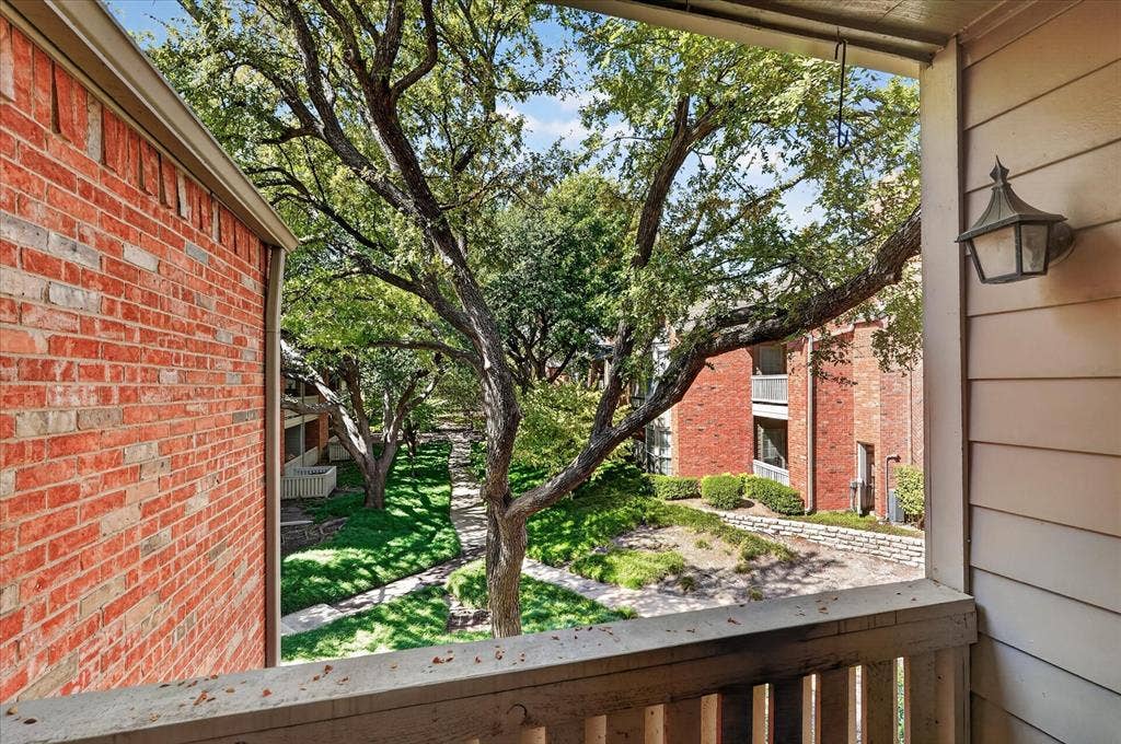 12660 Hillcrest Road, #5204, Dallas, TX 75230 Balcony with wood railing overlooking a lush courtyard, featuring mature trees and brick building exteriors