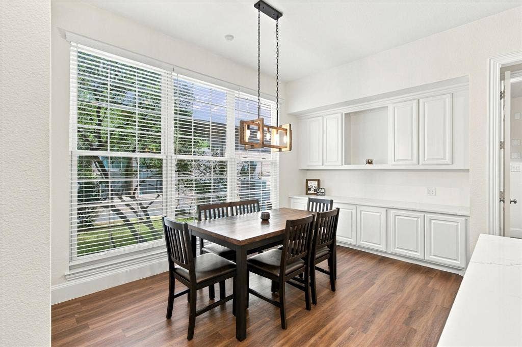 Wood-finish flooring, large multi-pane window with blinds, built-in cabinetry with white shaker-style doors, and a decorative chandelier