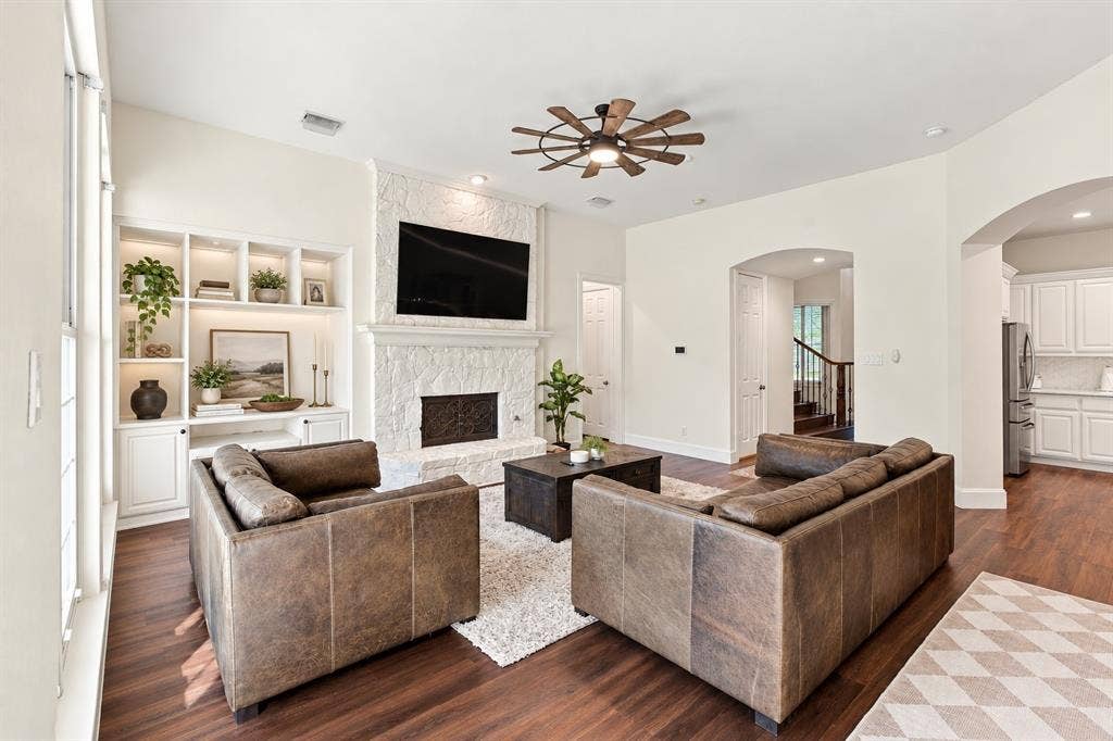 Living area featuring wood-finish flooring, white built-in shelving with integrated lighting, a stone fireplace, and an arched entryway