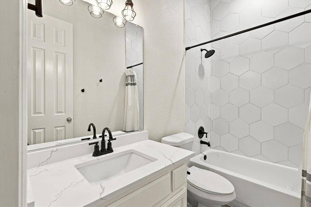 White hexagonal tile shower surround, matte black shower fixtures, white vanity with integrated rectangular sink, quartz-finish countertop, and matte black faucet