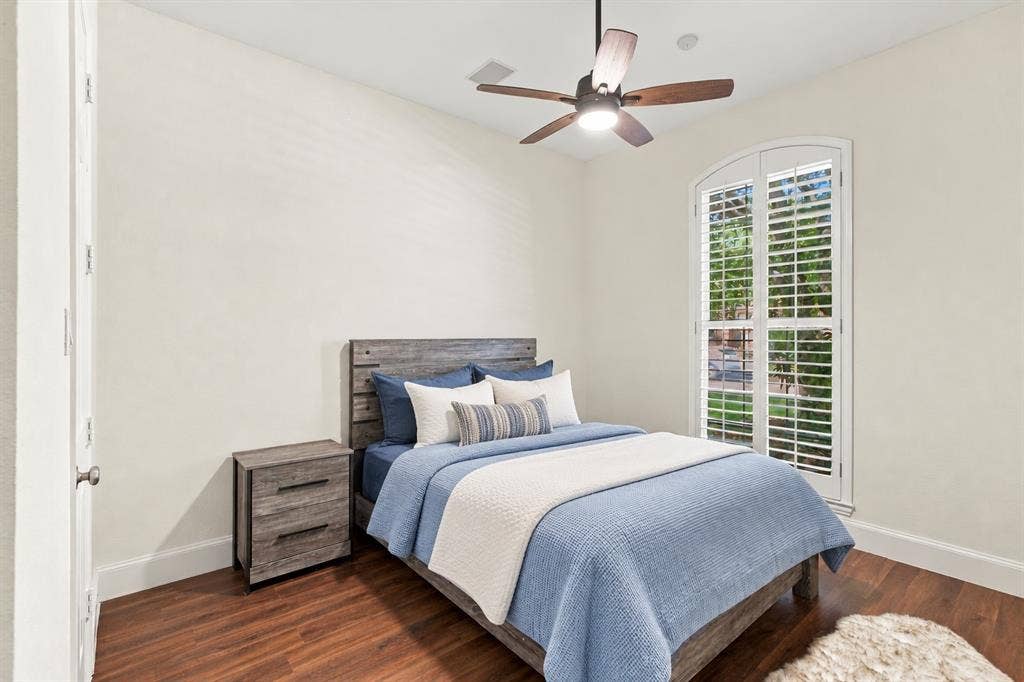 Wood-finish flooring, architectural arched window, white plantation shutters, and a ceiling fan with integrated lighting