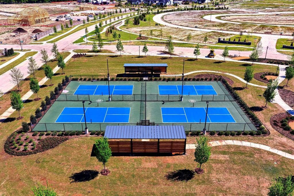 Four dedicated courts with blue playing surfaces, two covered seating areas, surrounding green landscaping, and paved walking paths