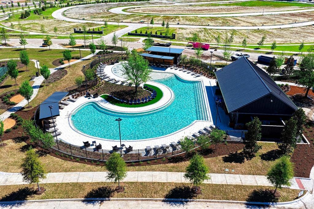 Expansive community pool with a surrounding paved deck, adjacent shaded lounge areas, and a modern pavilion with a dark-pitched roof
