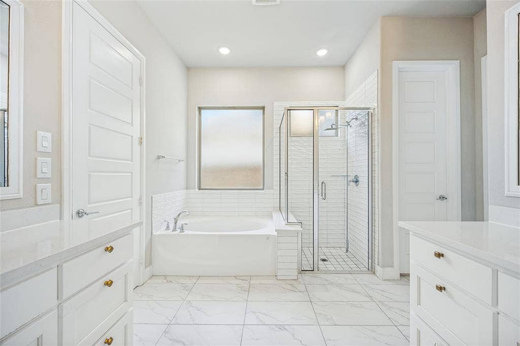 Spacious bathroom featuring a built-in bathtub, a walk-in shower with glass enclosure, dual vanities with white cabinetry, and large format floor tiling