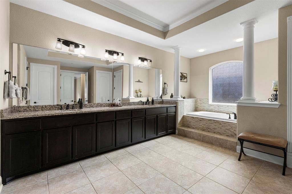 Double vanity with stone countertop, dark wood-finish cabinetry, built-in soaking tub with tile surround, arched window, and decorative architectural columns