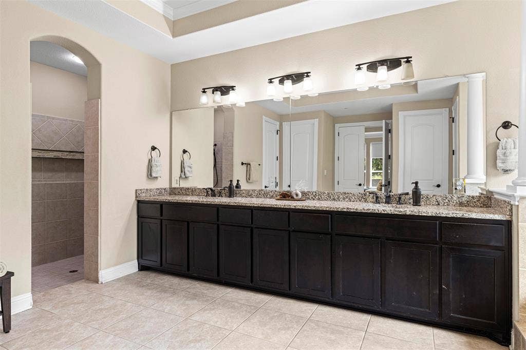 Dual-sink vanity featuring dark-stained cabinetry, expansive stone countertop, triple vanity lighting, and full-width wall mirror, complemented by tile flooring and tiled walk-in shower area