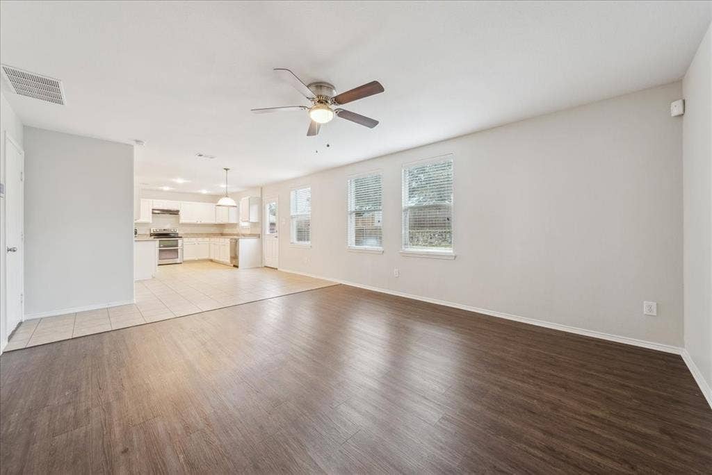 Wood-finish flooring, white walls, ceiling fan, multiple windows, and a partially visible kitchen with white cabinetry
