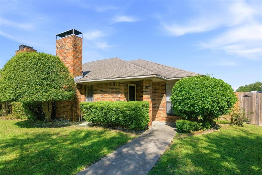 Brick exterior, twin brick chimneys, concrete walkway, pitched roof, and mature landscaping