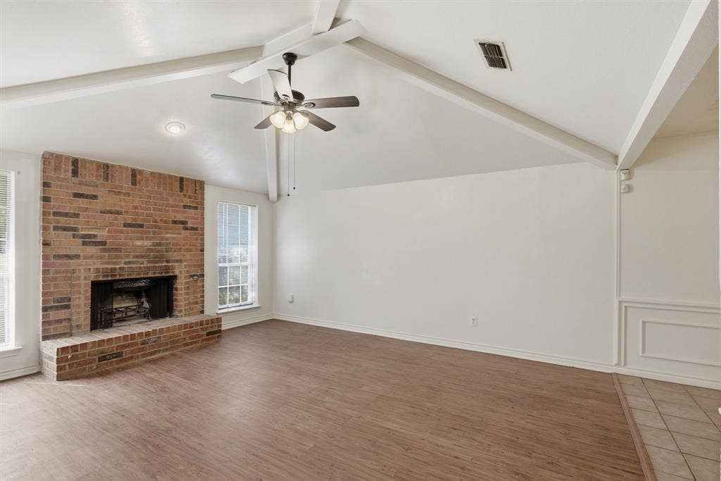 Vaulted ceiling with exposed beams, brick fireplace, wood-finish flooring, and a ceiling fan with integrated lighting