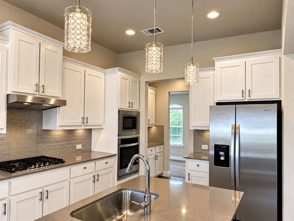 Chef's kitchen featuring white cabinetry with brushed nickel hardware, stainless steel appliances including a side-by-side refrigerator and dual wall ovens, quartz countertops, a gas cooktop with stainless steel hood, subway tile backsplash, and a large center island with undermount sink and chrome faucet