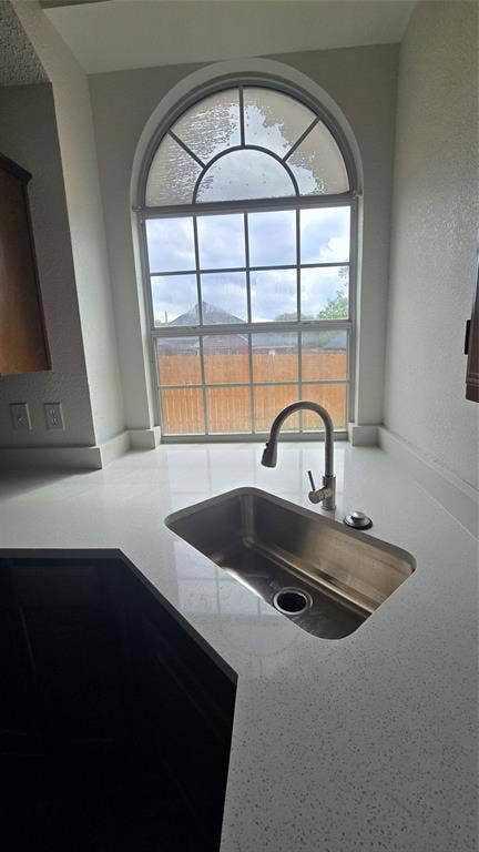 Arched window with divided lites, undermount stainless steel sink, gooseneck faucet, light-toned countertop, and dark base cabinetry