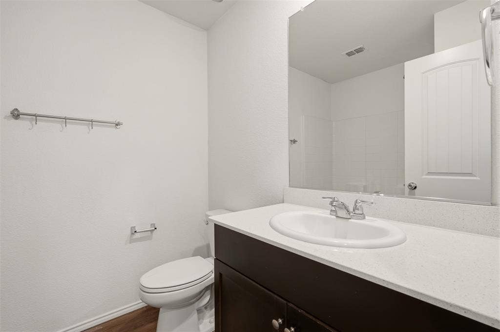 Wood-finish flooring, dark wood vanity with white countertop, integrated sink with chrome faucet, large wall-mounted mirror, and white toilet
