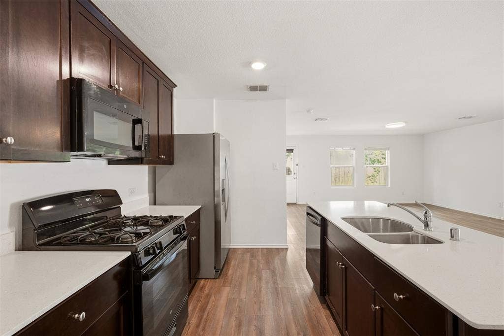 Kitchen featuring wood-finish flooring, dark wood cabinetry, light countertops, stainless steel appliances, and a kitchen island with an undermount double sink