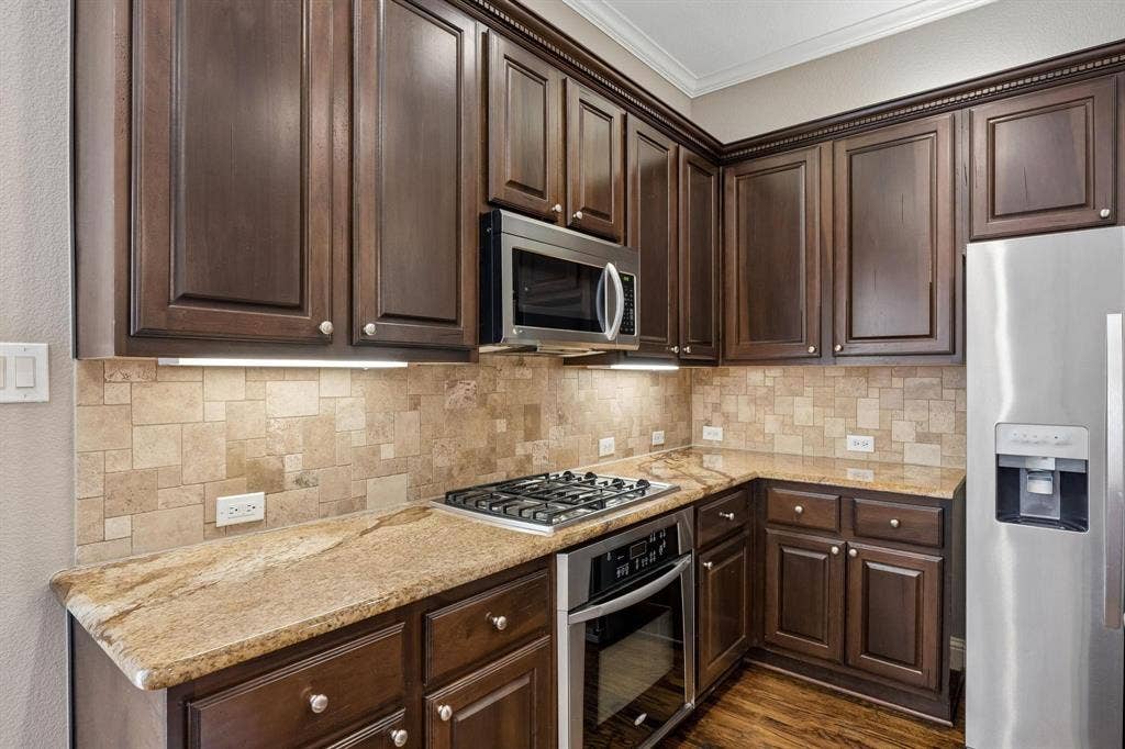 Dark wood-finish cabinetry with crown molding, stainless steel appliances, stone slab countertops, a gas range with oven, and a tiled backsplash