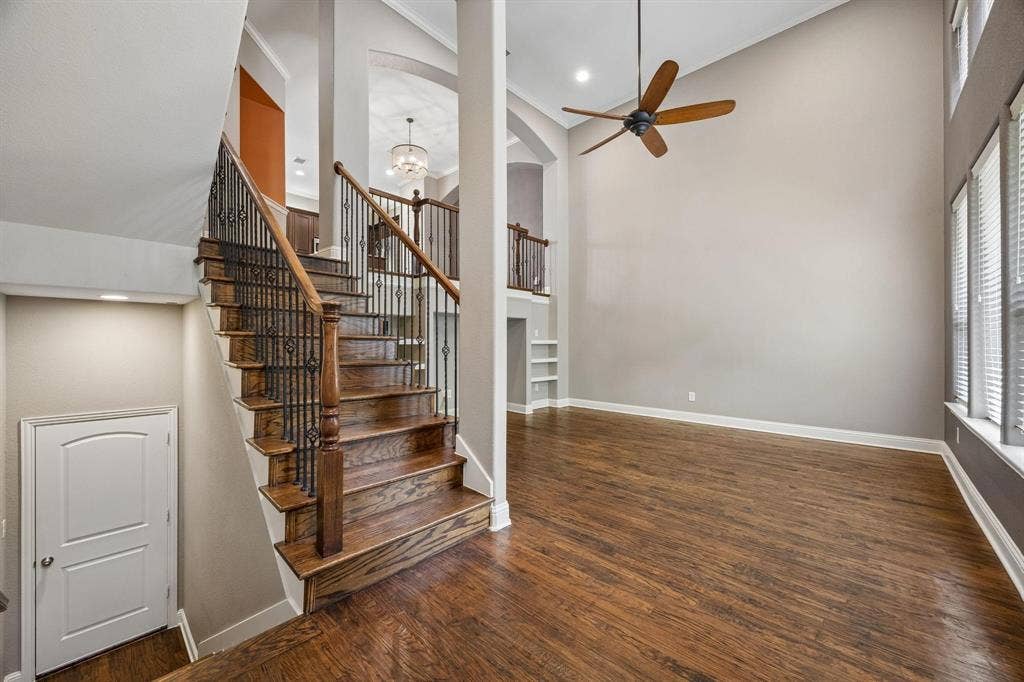 Hardwood flooring throughout, a grand staircase with wrought iron balusters, high ceilings, built-in shelving, and multiple windows with blinds