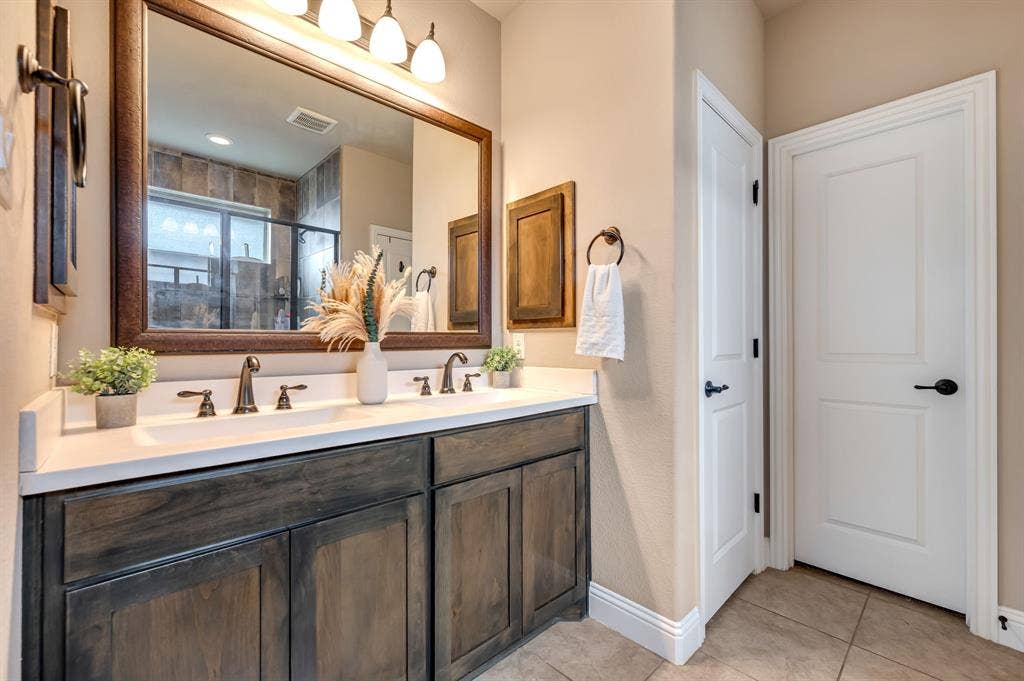 Double-sink vanity featuring stained wood cabinetry, white countertop, dark metal hardware, and a large framed mirror with overhead lighting