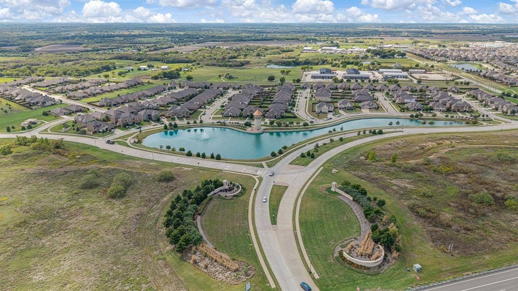 Aerial perspective of a residential community featuring a central pond with a fountain, landscaped entrance signage, and paved street infrastructure