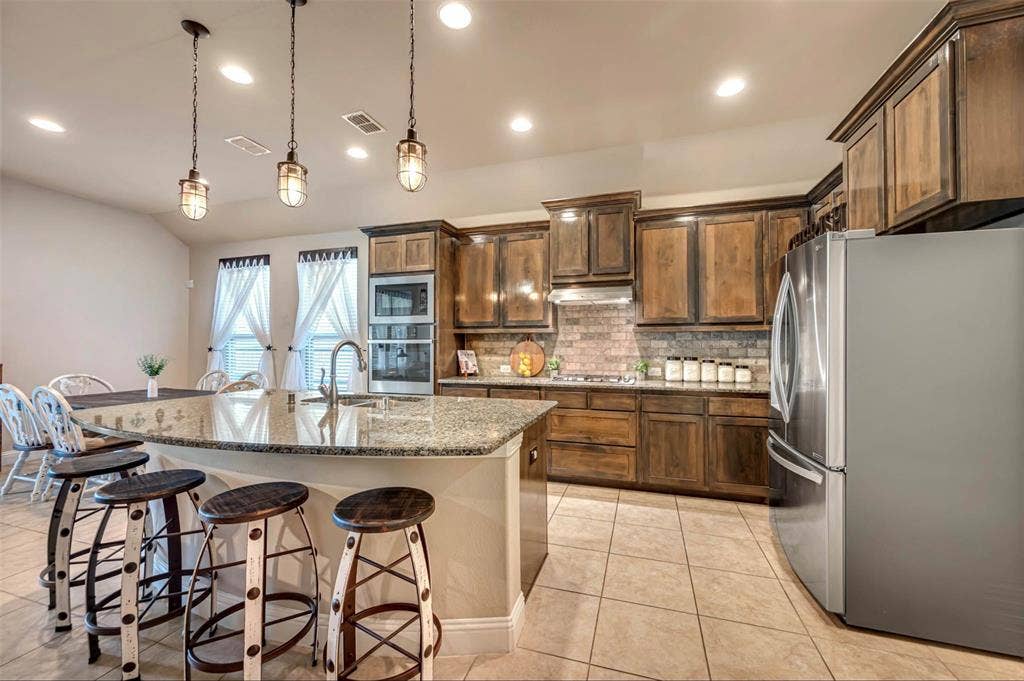 Kitchen featuring stained wood cabinetry, stone-surface island with bar seating, brick-style tile backsplash, stainless steel built-in oven, and pendant lighting