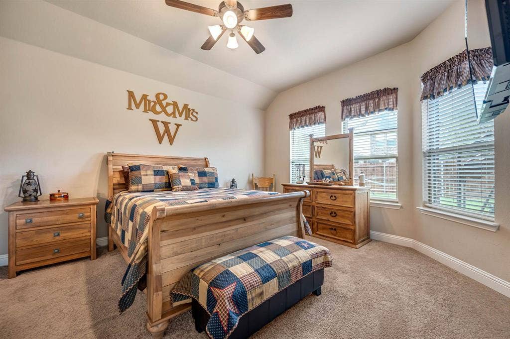 Neutral-toned interior room featuring vaulted ceilings, carpeted flooring, a multi-blade ceiling fan with integrated lighting, and multiple windows with horizontal blinds