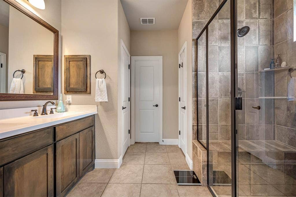 Bathroom featuring stained wood cabinetry, white countertop with dual under-mount sinks, tile flooring, and a walk-in shower enclosed by a framed glass door with wall-tile surround