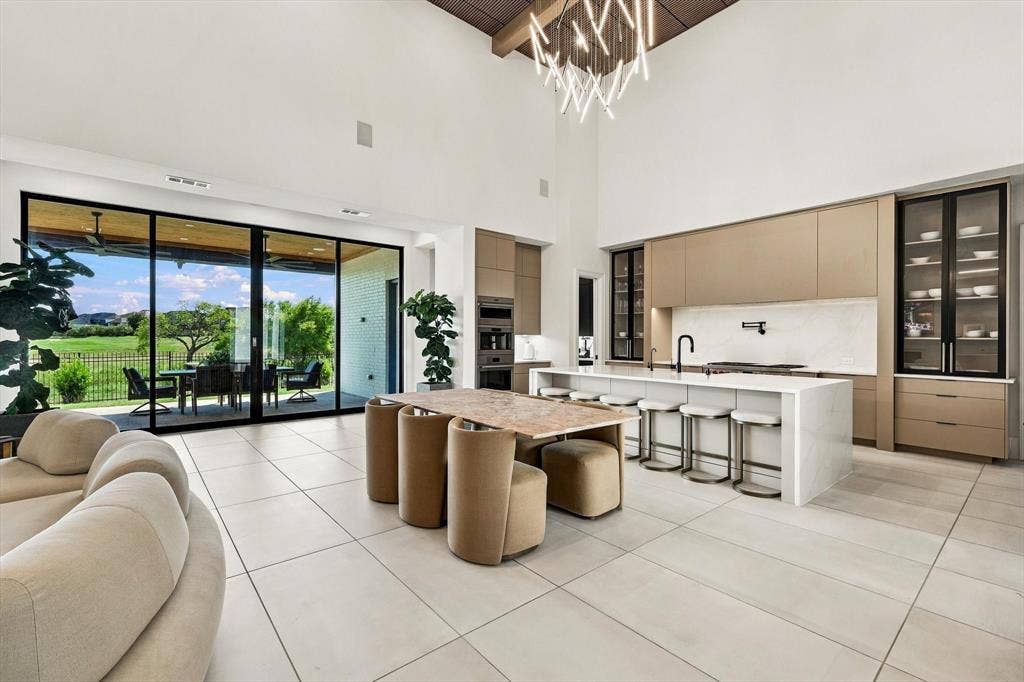 4301 Corabelle Lane, Prosper, TX 75078 Expansive main living area featuring high vaulted ceilings, large format tile flooring, a modern kitchen with a waterfall island, and floor-to-ceiling glass panel doors opening to an outdoor patio