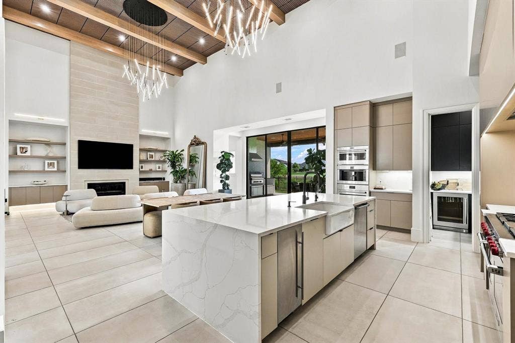 4301 Corabelle Lane, Prosper, TX 75078 Two-story ceiling with exposed wood beams and contemporary chandeliers, stone fireplace with built-in shelving, expansive kitchen island with a farmhouse sink, integrated stainless steel appliances, and large format tile flooring