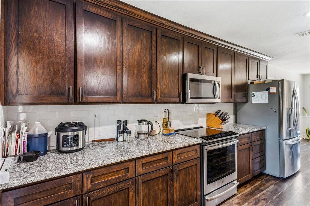 Dark wood cabinetry, speckled countertops, stainless steel appliances, white subway tile backsplash, wood-finish flooring