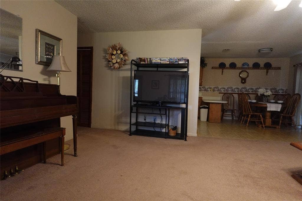 Carpeted living area with textured ceiling, leading to a dining area featuring wood-finish flooring