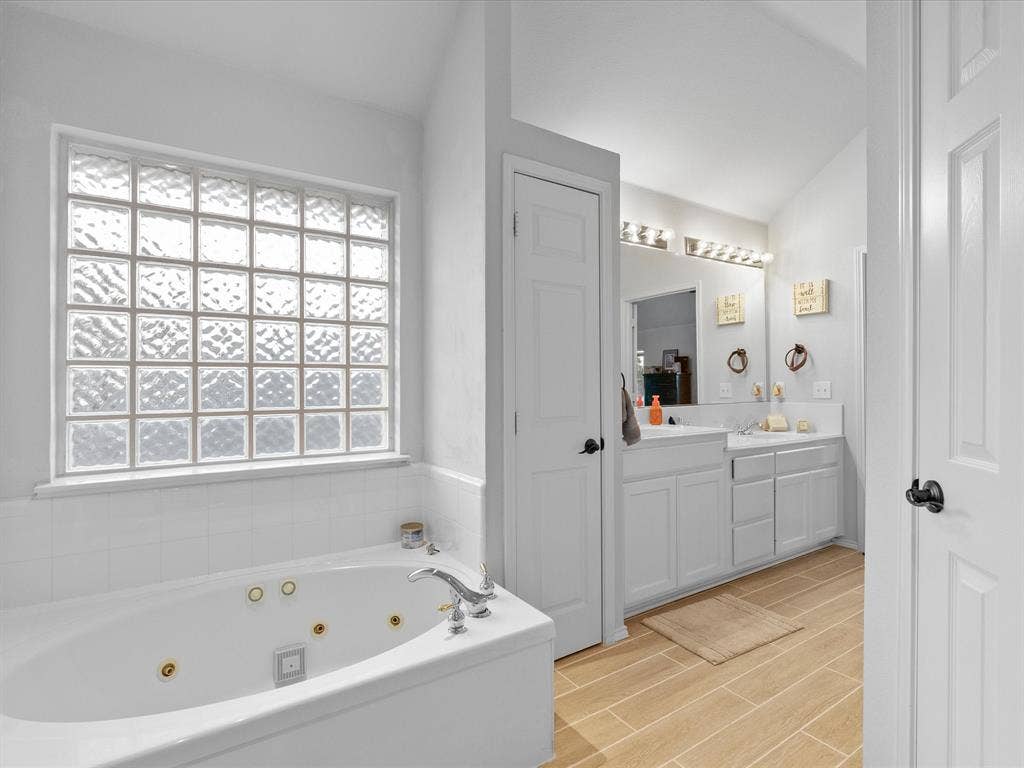 3303 Felicia Court, McKinney, TX 75070 Jetted soaking tub, glass block window, wood-finish tile flooring, dual-sink vanity with white cabinetry, and multi-light vanity fixtures