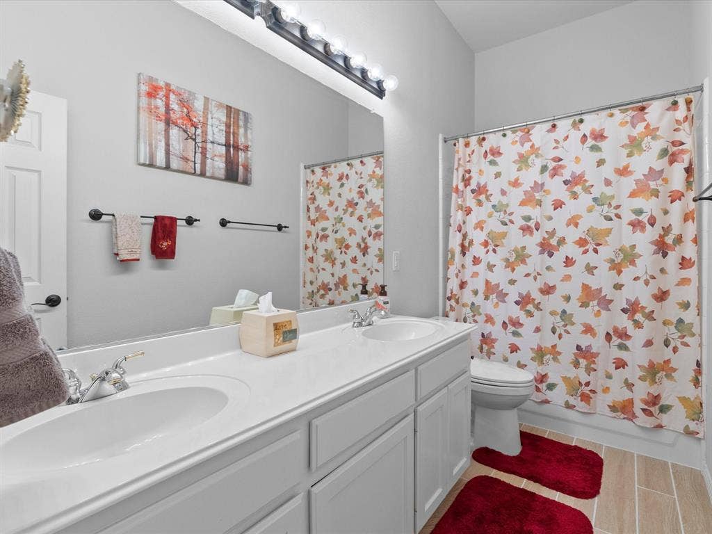 3303 Felicia Court, McKinney, TX 75070 Dual-sink vanity with a solid-surface countertop, full-width mirror, five-bulb integrated light fixture, and wood-finish tile flooring