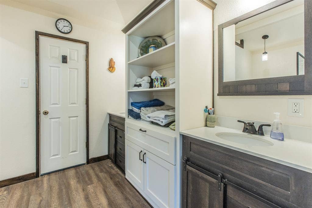 Wood-finish flooring, white paneled door with dark trim, built-in shelving, vanity with white countertop and integrated sink, and dark wood-frame mirror