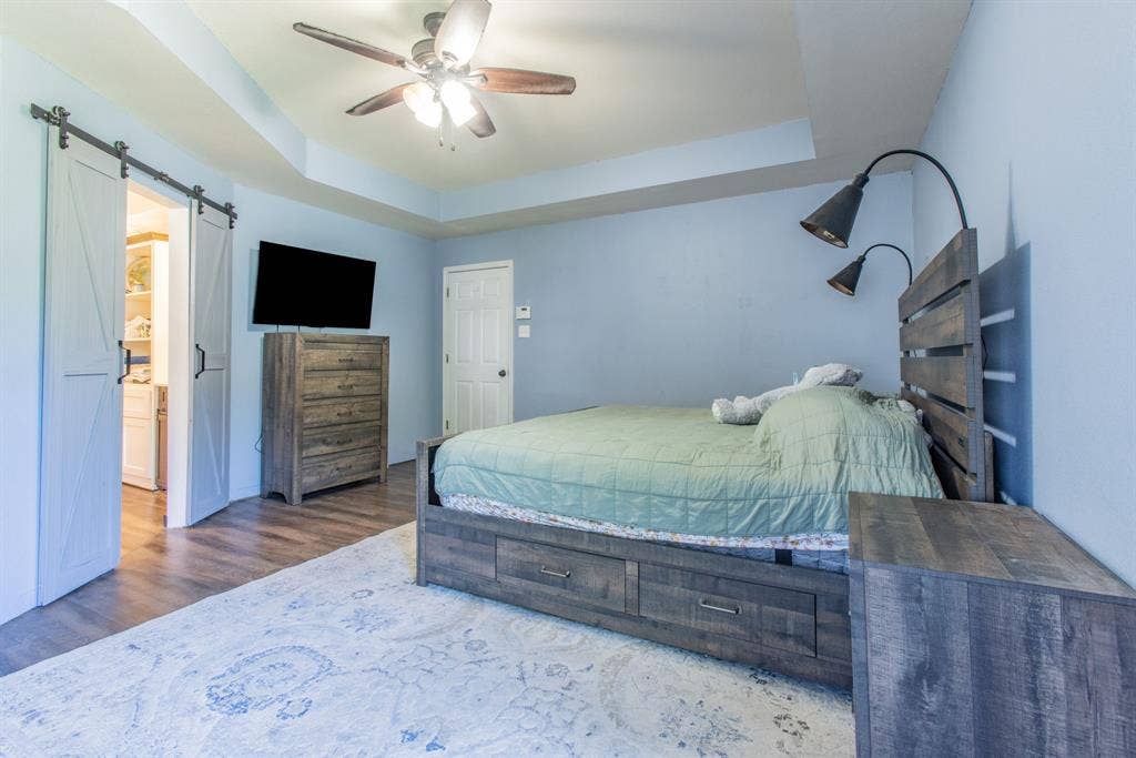 Tray ceiling with ceiling fan, wood-finish flooring, sliding barn doors, and attached wall-mounted lighting fixtures