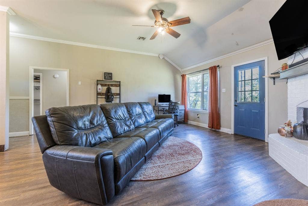 Vaulted ceiling, wood-finish flooring, white brick fireplace, ceiling fan, and a blue paneled door with glass inserts